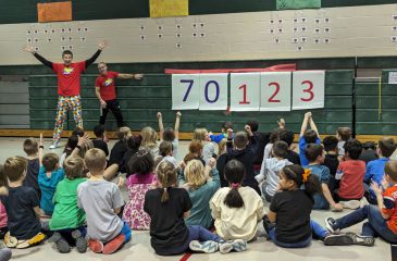 Large group of elementary aged kids sitting cross-cross on the gym floor, celebrating the reveal of $70,123 raised for AHA